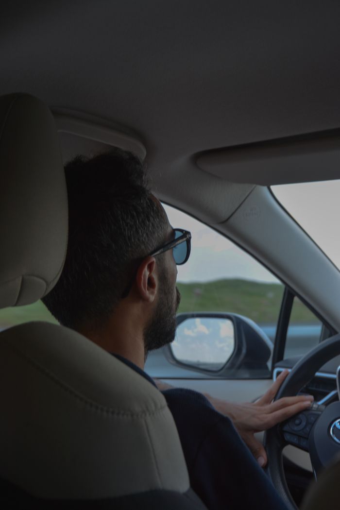 Side view of a young man wearing sunglasses driving a car on a scenic road, relaxed and focused.